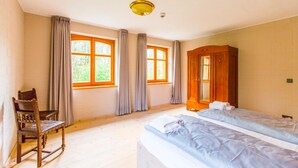 Una habitación con dos camas, un armario de madera, una silla y ventanas grandes con cortinas.