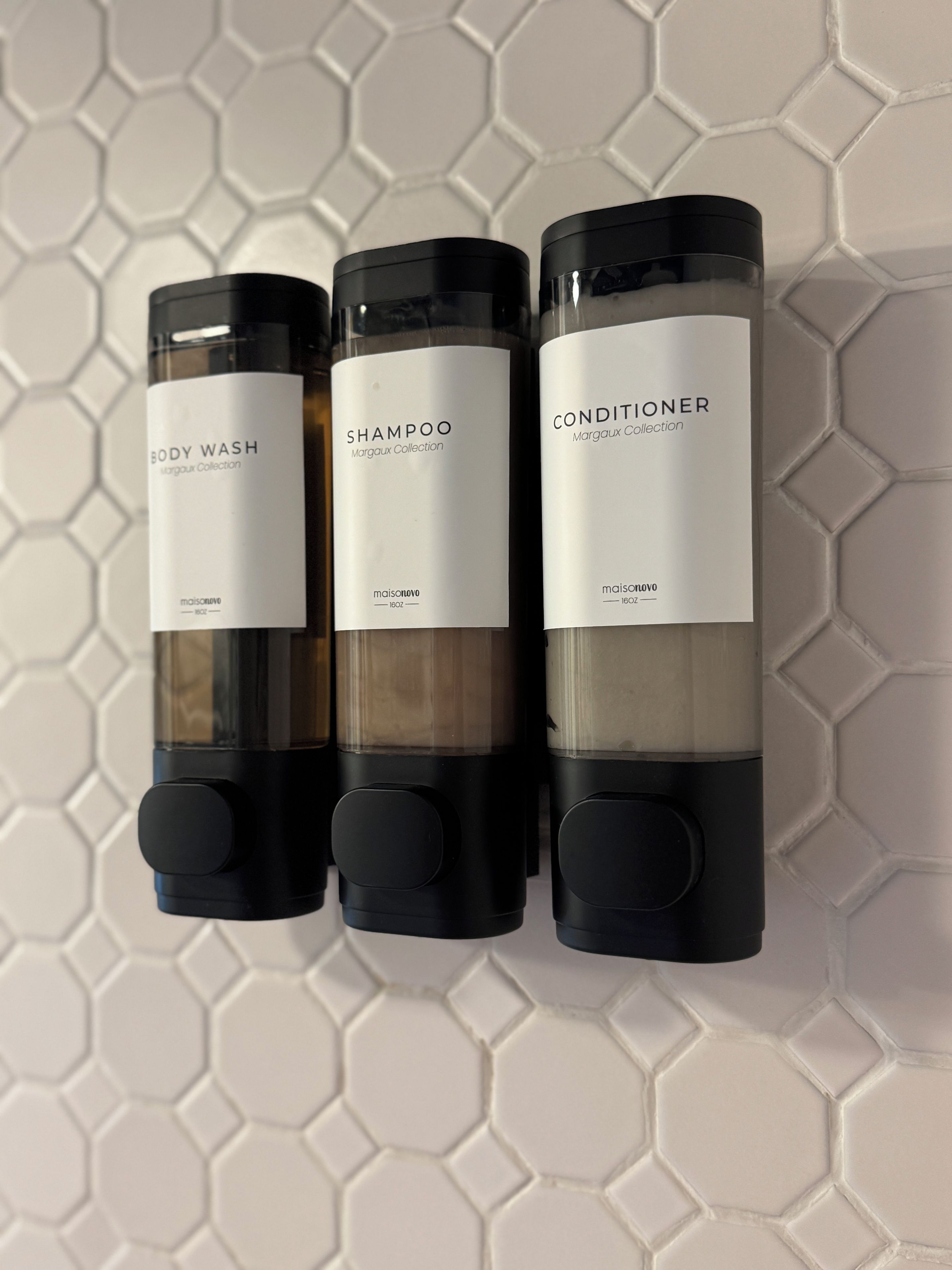 Tres botellas de gel de baño, champú y acondicionador de la colección Margaux, dispuestas verticalmente sobre una pared con motivo hexagonal.