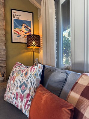 A room with a sofa, patterned cushions, a lamp, a framed picture on the wall, and a window with curtains. - Big Sky Ski & Snowboard Escape, Cozy Fireside Nights. Black Otter 55 Basecamp. (Big Sky)