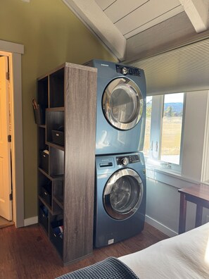 A laundry room with a Samsung washer and dryer, wooden shelving, and a window with a view. - Big Sky Ski & Snowboard Escape, Cozy Fireside Nights. Black Otter 55 Basecamp. (Big Sky)