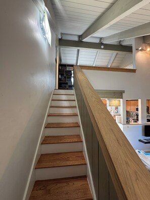 A staircase with wooden steps and a wooden handrail leading to a second-floor area with a kitchen and living space. - Big Sky Ski & Snowboard Escape, Cozy Fireside Nights. Black Otter 55 Basecamp. (Big Sky)