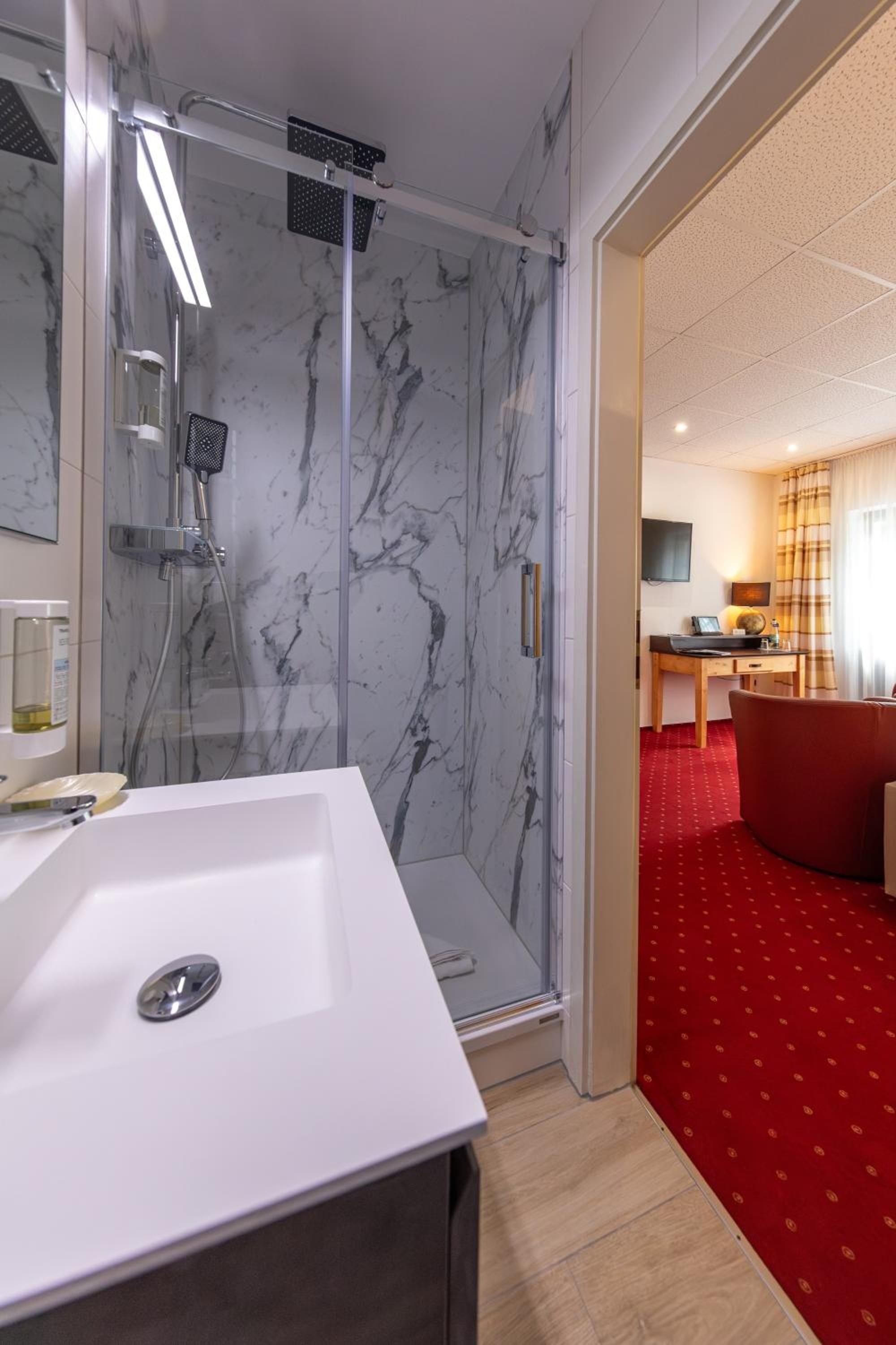 Ein modernes Badezimmer mit einer Duschkabine aus Marmor, einem weißen Waschbecken und einem roten Teppichbereich, der in einen Raum mit Schreibtisch und Fernseher führt.