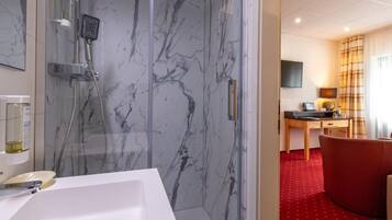 Ein modernes Badezimmer mit einer Duschkabine aus Marmor, einem weißen Waschbecken und einem roten Teppichbereich, der in einen Raum mit Schreibtisch und Fernseher führt.