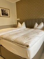 Cama bem arrumada com um edredom branco texturizado e almofadas, cabeceira acolchoada e papel de parede estampado ao fundo.