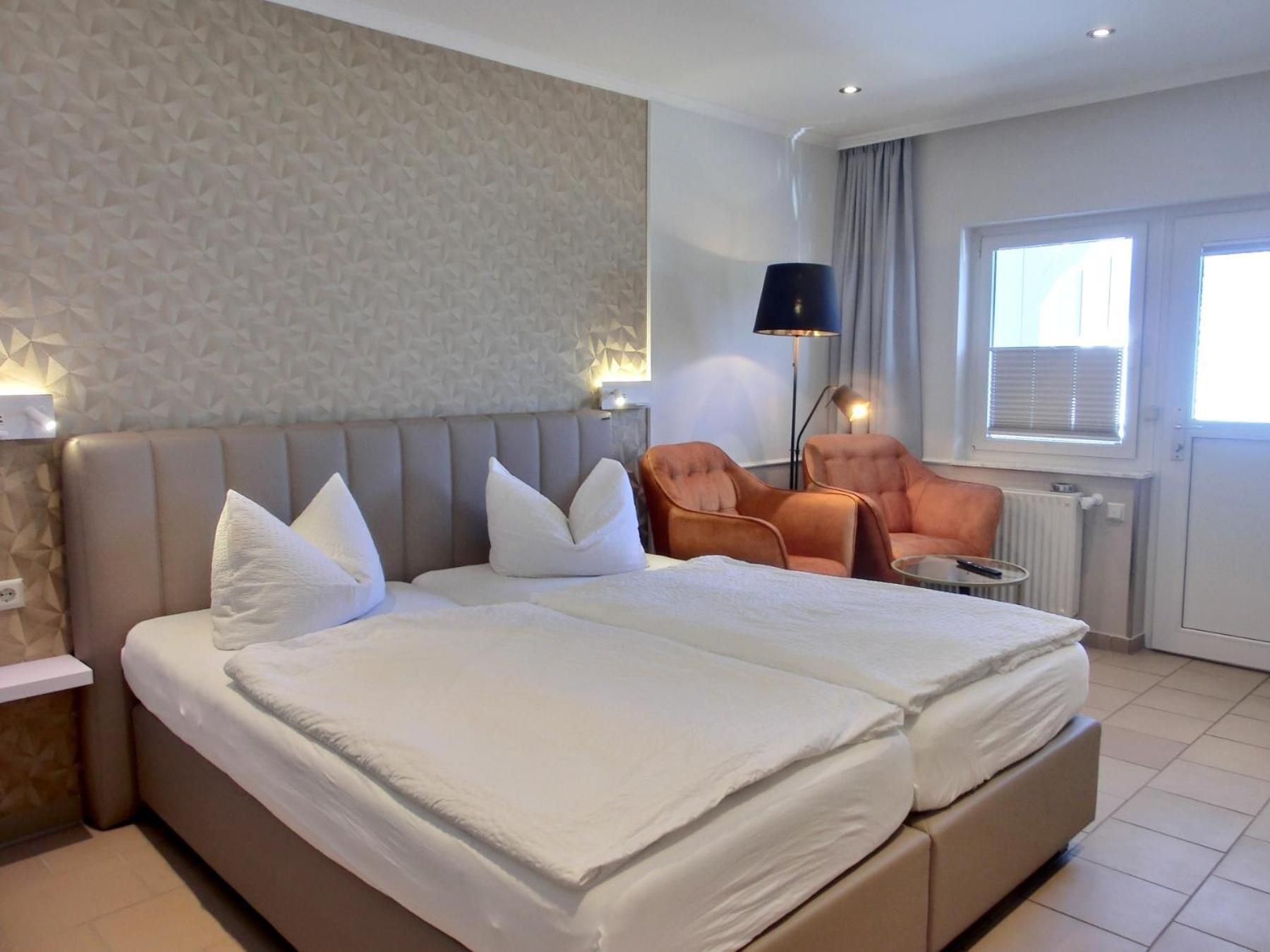 Una habitación de hotel con una cama grande, dos sillones, una lámpara de pie y un ventanal con persianas.