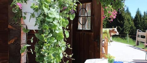 A wooden cabin with a porch, a hanging planter with green foliage, a round table with a chair, and a decorative cow statue.