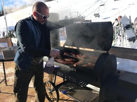 Una persona asando carne al aire libre con una parrilla portátil sobre ruedas.