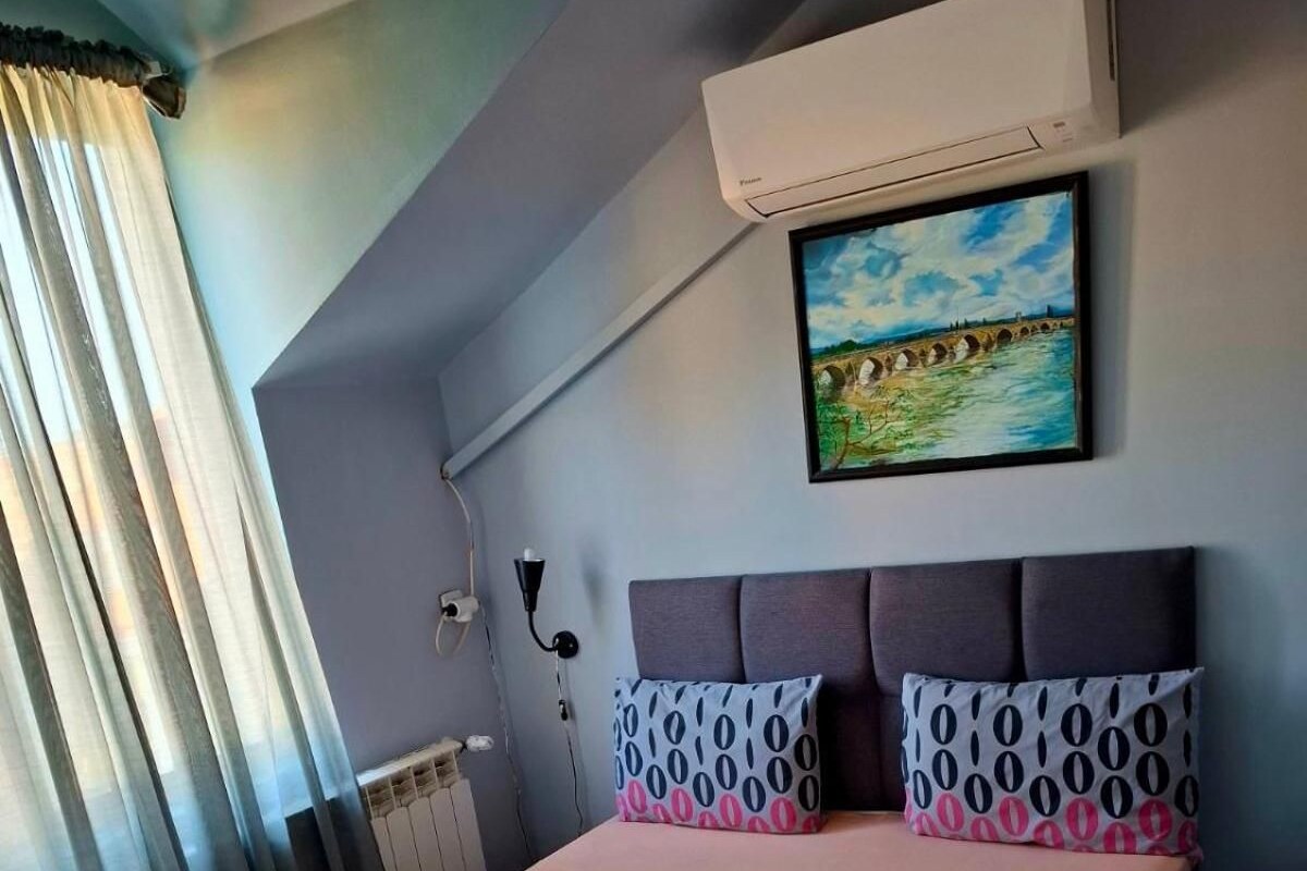 A bedroom with a bed, a painting, an air conditioner, and a window with curtains.