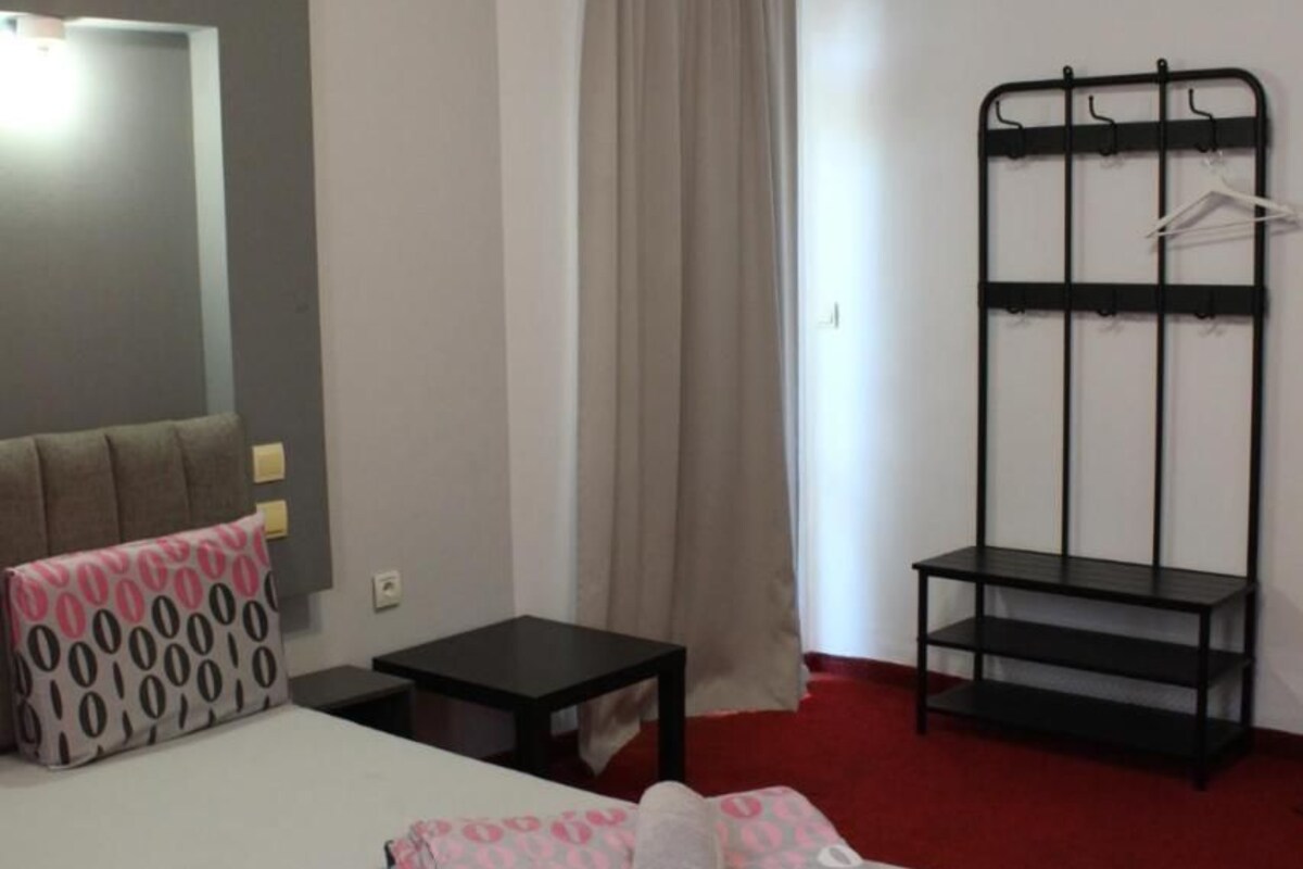 A hotel room with a bed, a bedside table, a hanging rack, and a window with curtains.