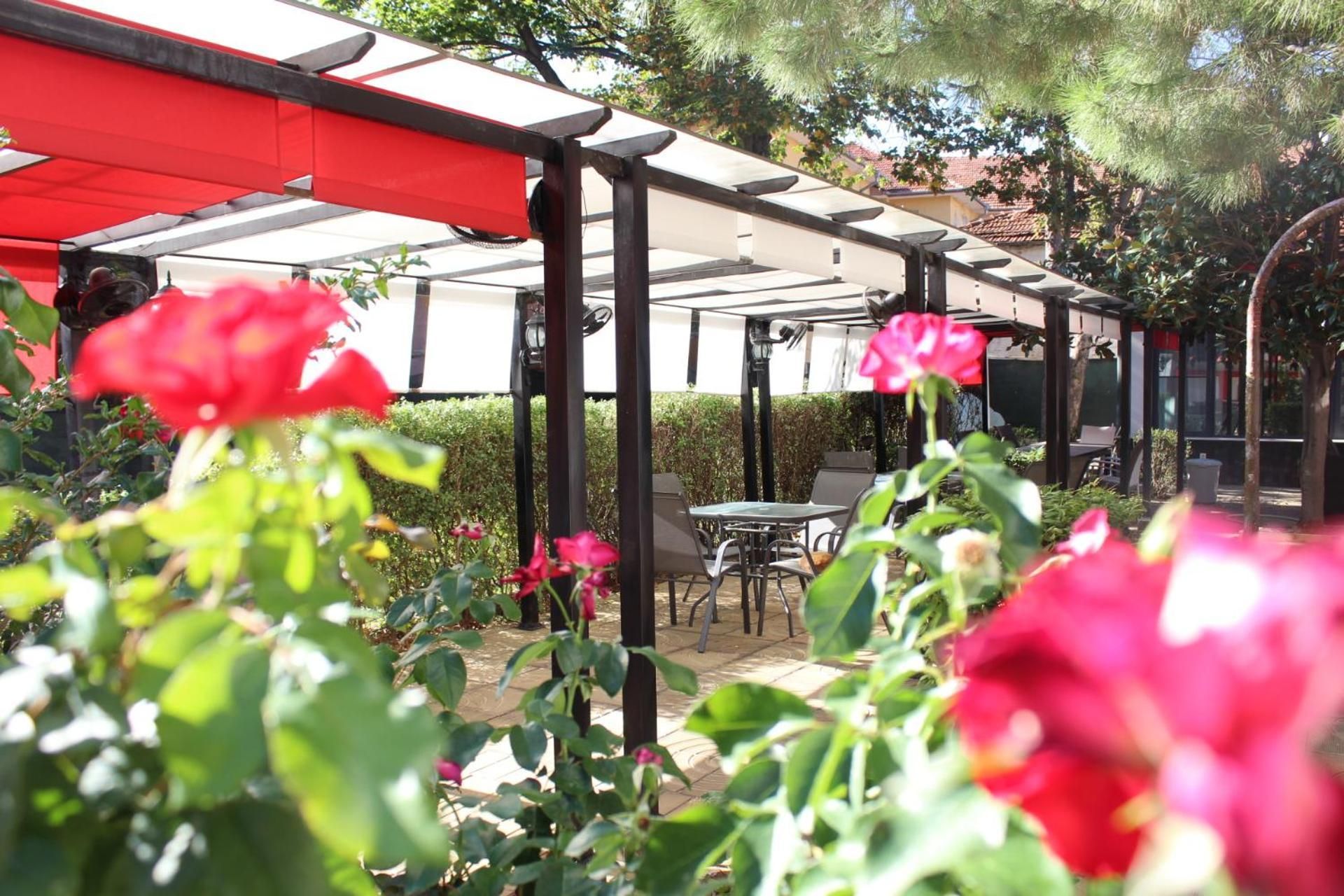A covered outdoor seating area with tables and chairs, surrounded by greenery and flowering plants.
