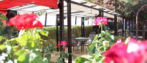 A covered outdoor seating area with tables and chairs, surrounded by greenery and flowering plants.