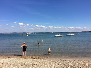 En strand med småsten, med båtar på vattnet och människor på avstånd.