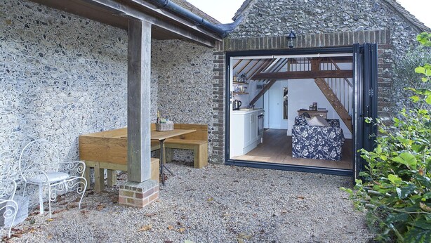 A stone exterior with a wooden bench and a table, a doorway leading to a room with a kitchen, and a staircase visible through the doorway. - Shepherds Barn (Chichester)