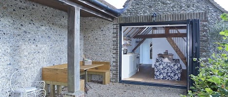 A stone exterior with a wooden bench and a table, a doorway leading to a room with a kitchen, and a staircase visible through the doorway.