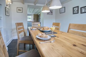 A dining area with a wooden table, chairs, and a breakfast setting including croissants and juice. - Dollars Cottage 4 Bed Llangennith (Swansea)