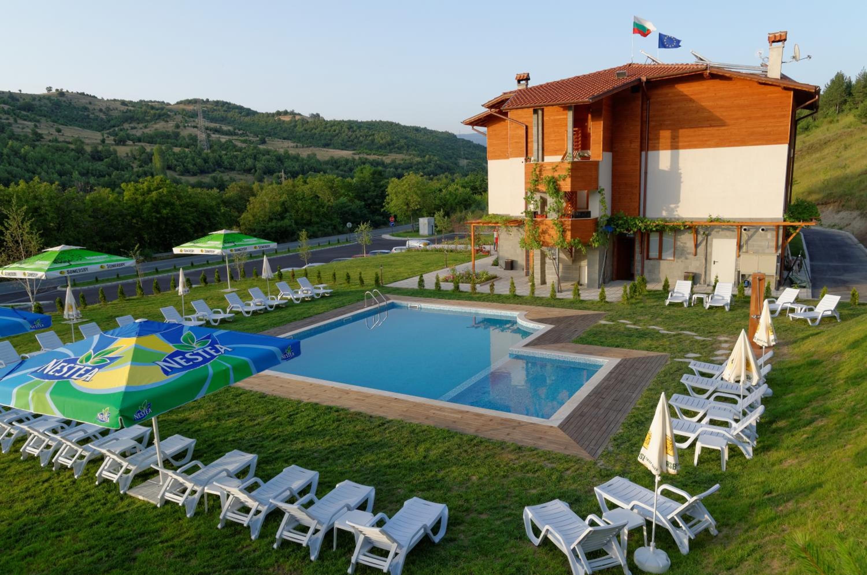 Zona de piscina con sillas de descanso y sombrillas, un edificio con una bandera y un paisaje montañoso al fondo.