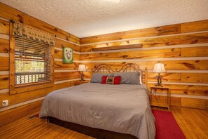 A wooden cabin bedroom with a bed, two bedside tables, a window with blinds, and a wall-mounted lamp. - Cozy Bears Cabin (Pigeon Forge)