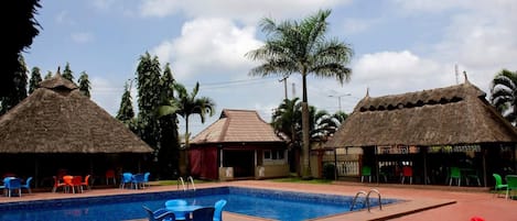 A swimming pool with blue chairs and tables, surrounded by buildings with thatched roofs and palm trees.