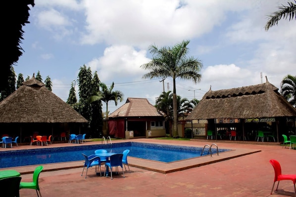 A swimming pool with blue chairs and tables, surrounded by buildings with thatched roofs and palm trees. - Villa Park Hotels (Lagos)