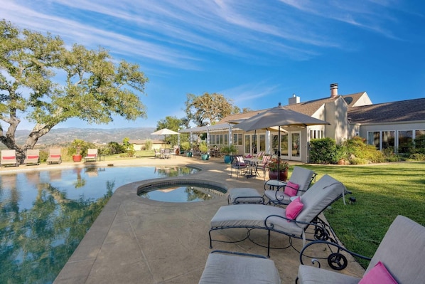 A pool area with lounge chairs and a hot tub, surrounded by a patio with outdoor seating and a view of a house.