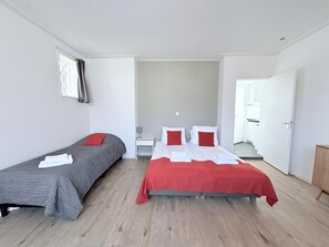 Quarto de hotel com duas camas de solteiro, uma mesinha de cabeceira com luminária e vista para o corredor.