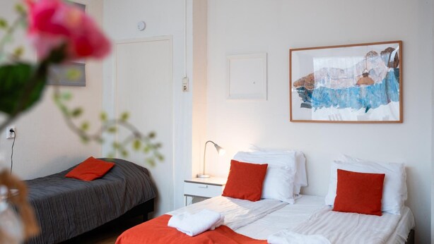 A hotel room with two beds, a nightstand, a lamp, and a painting on the wall. - Hotel Cafe De Harmonie (Edam)