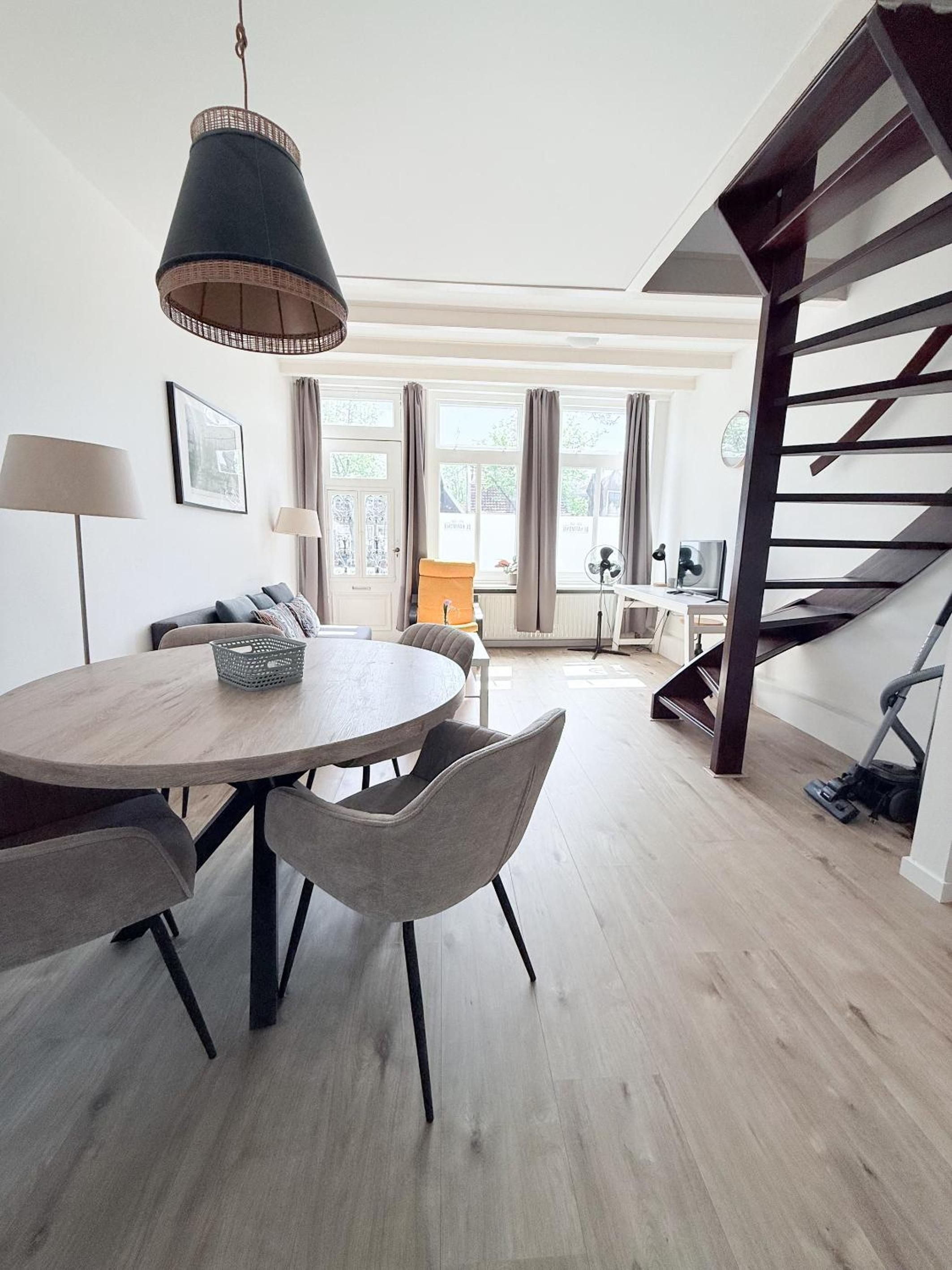A modern living room with a round wooden dining table, grey upholstered chairs, a hanging light fixture, a staircase, and a view of the outside through large windows.