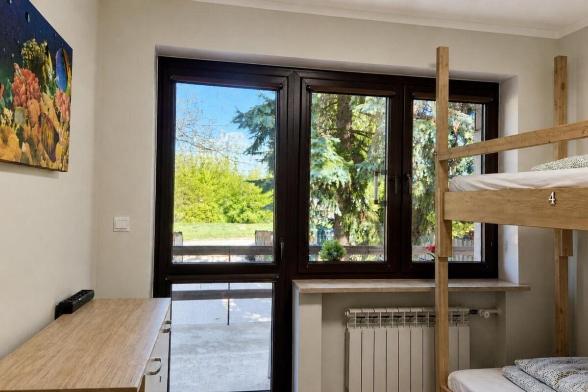 A room with a bunk bed, a desk, a radiator, and a large window with a view of greenery.