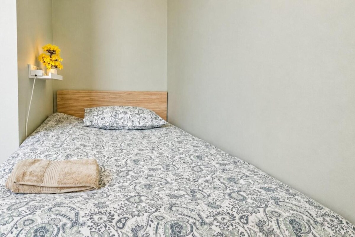 A single bed with a patterned bedspread, a wooden headboard, and a bedside lamp with a yellow flower.