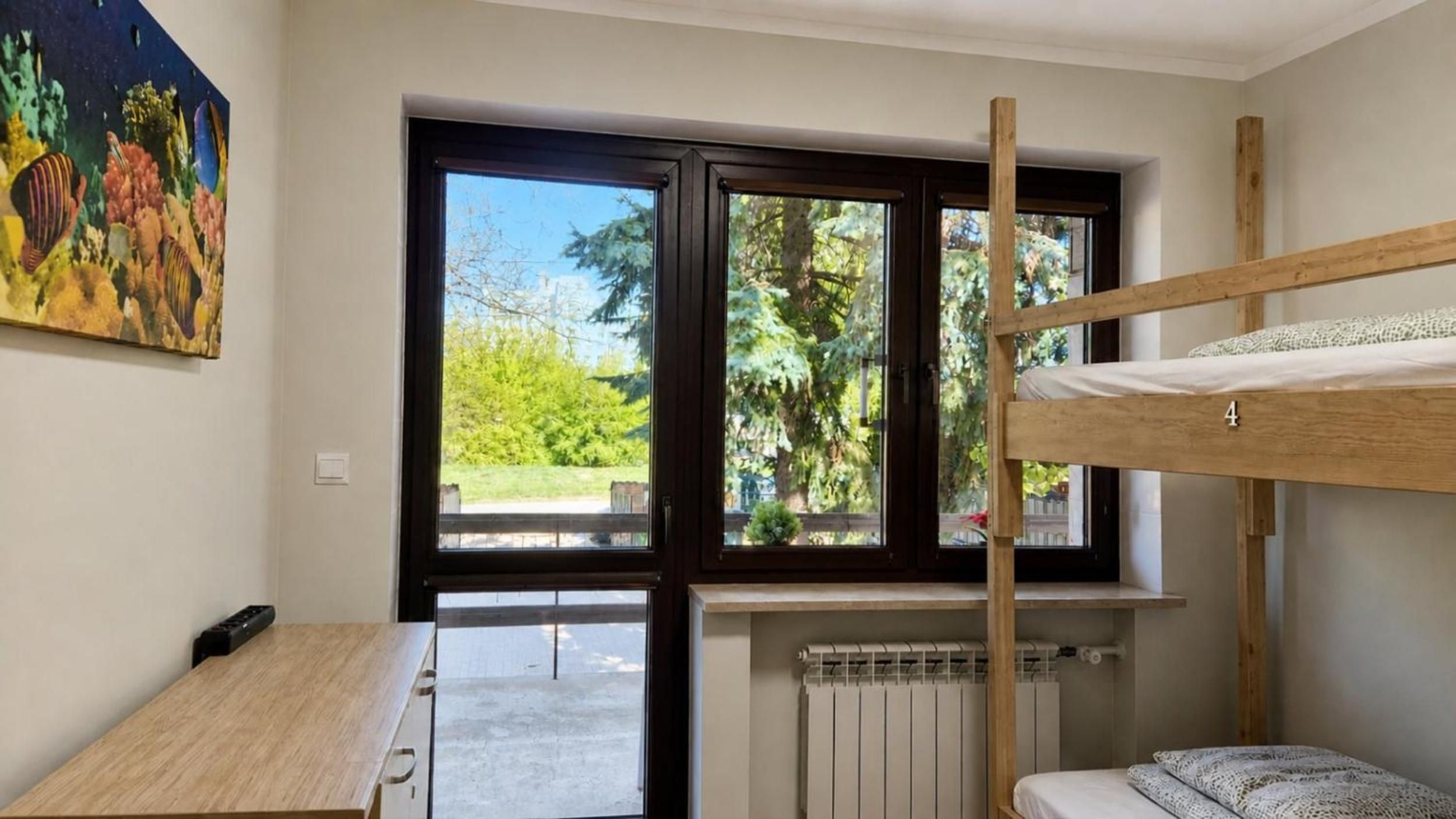 A room with a bunk bed, a desk, a radiator, and a large window with a view of greenery.