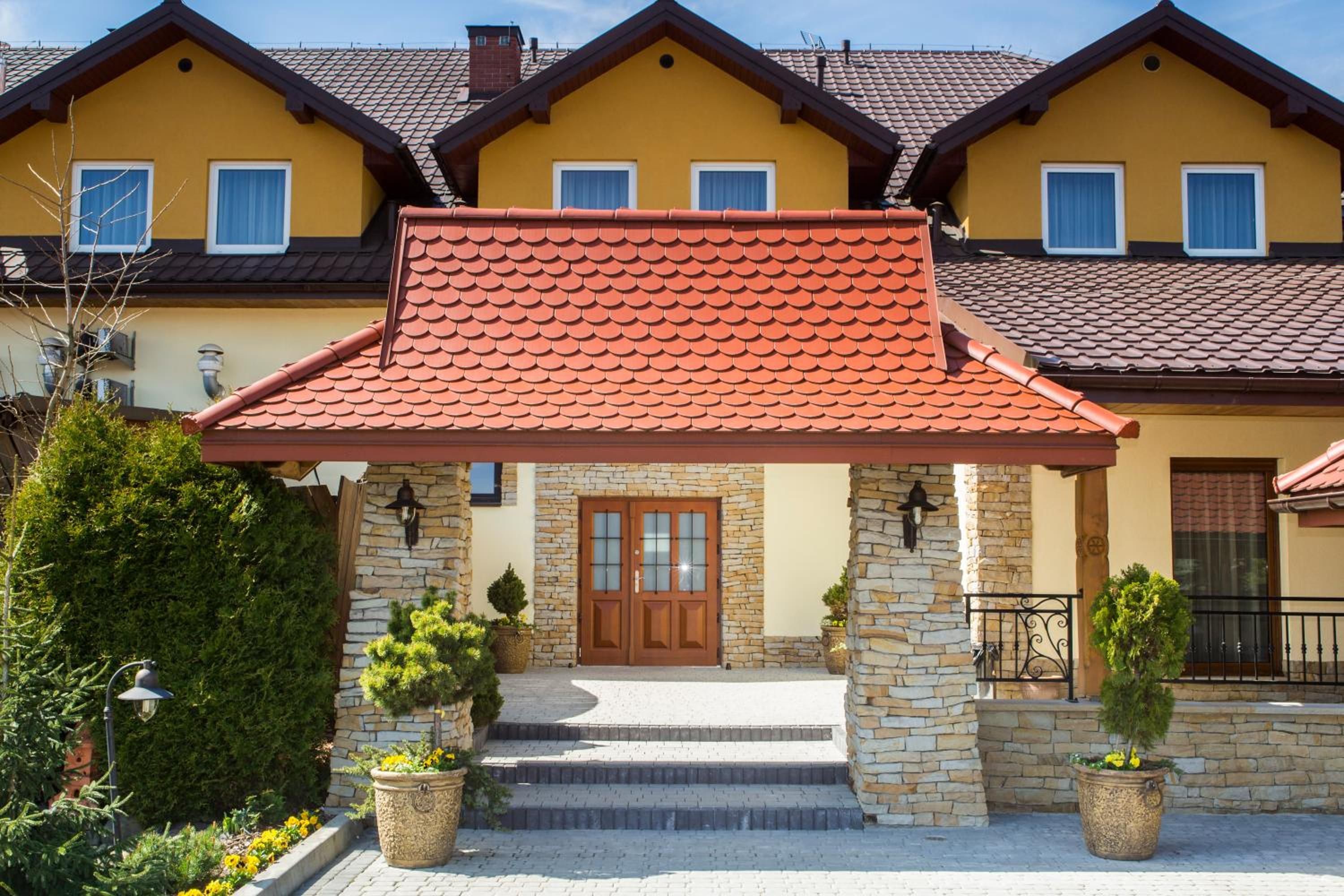 A two-story house with a red-tiled roof, stone pillars, and a wooden front door.