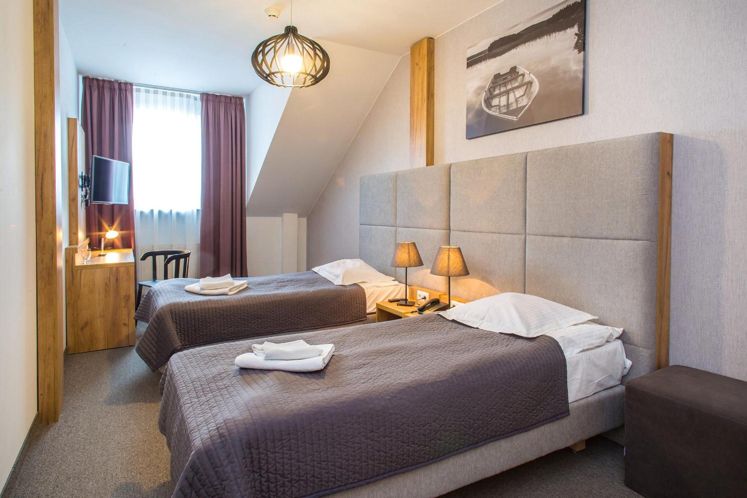 A hotel room with two beds, a large window with curtains, a mounted TV, and a painting on the wall.