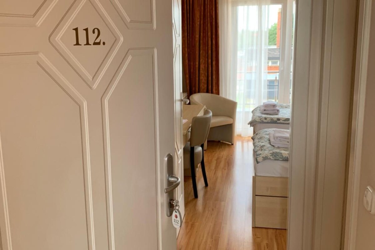 Habitación de hotel con una cama, una silla y un escritorio. El piso es de madera y la puerta tiene el número 112.