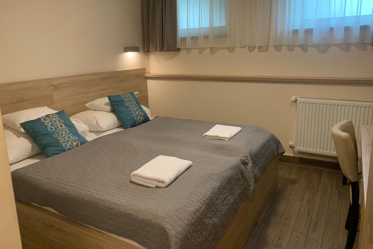 Quarto de hotel com uma cama de casal, duas almofadas, mesa de cabeceira, uma cadeira, janela com cortinas e radiador.