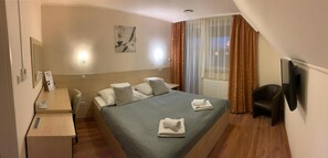 Quarto de hotel com cama, escrivaninha, cadeira, televisão e janela com cortinas.