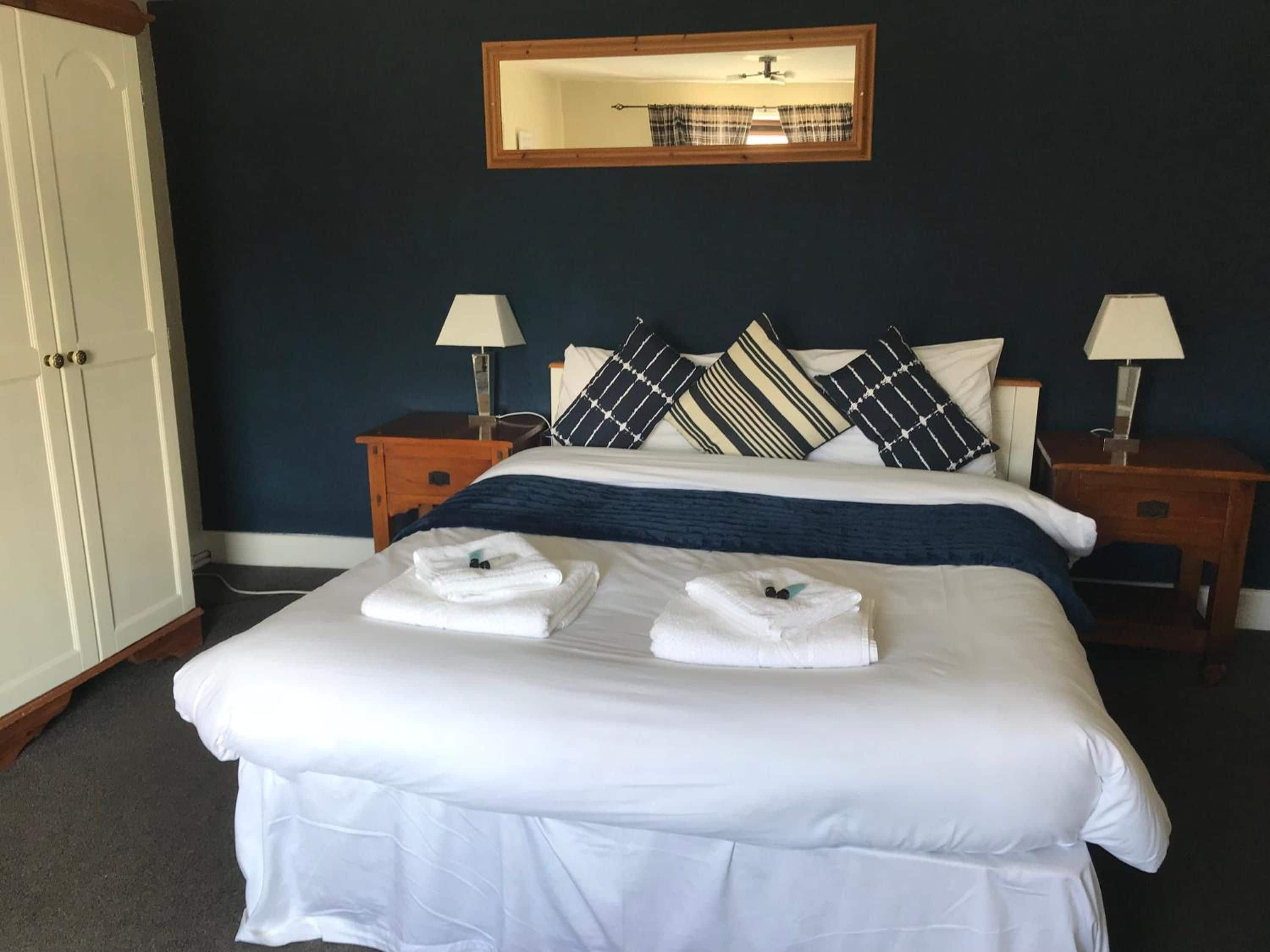 A neatly made bed with white linens and towels, a dark blue wall, a mirror, and two bedside tables with lamps.