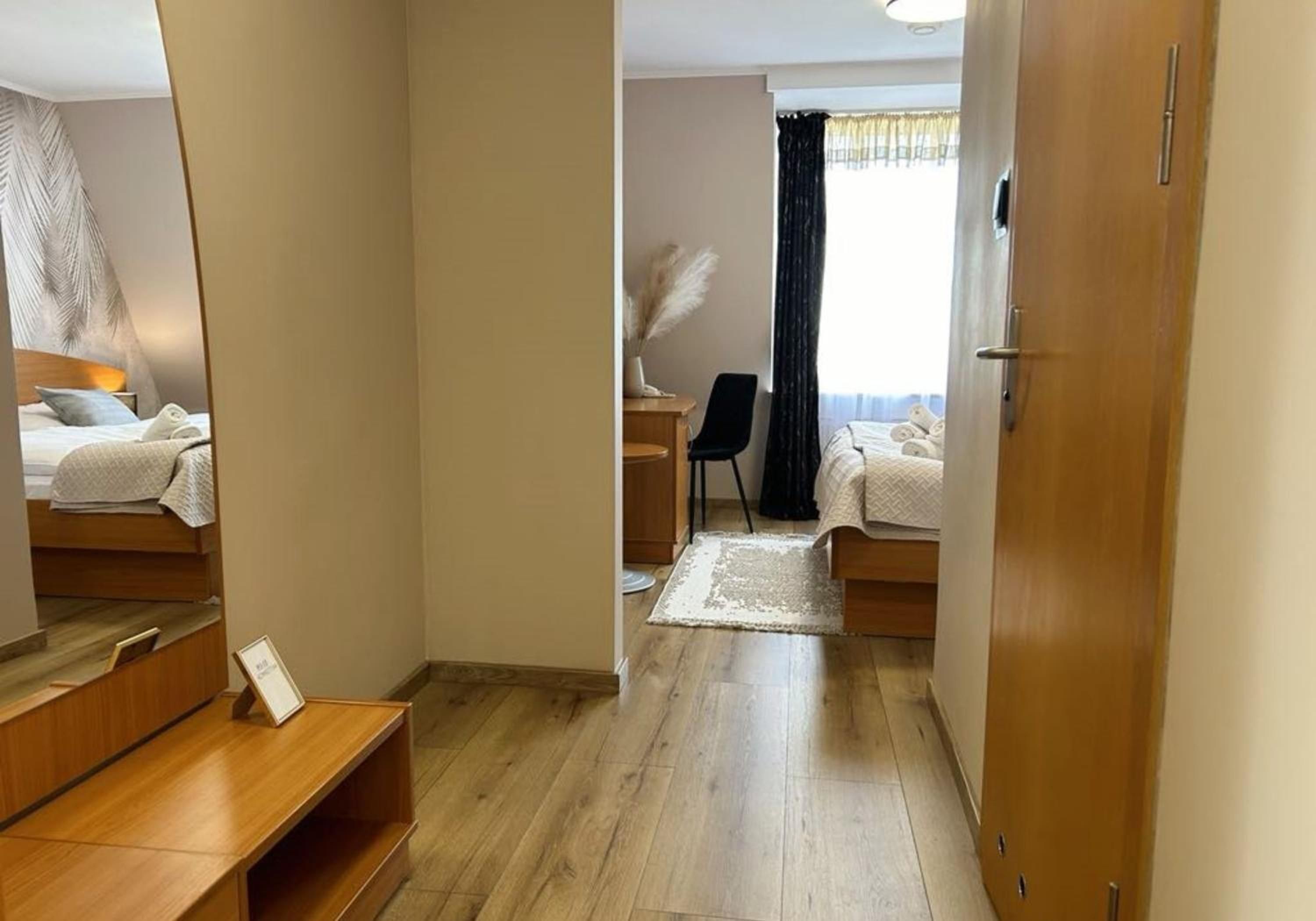A hotel room with a wooden floor, a bed with white bedding, a desk with a chair, and a window with curtains.