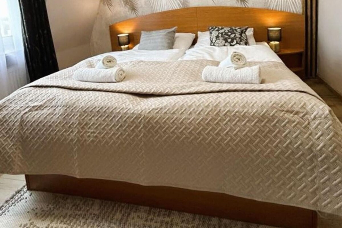 Habitación de hotel con una cama grande, cabecera con estampado de hojas, dos lámparas de noche y un tapete texturizado.