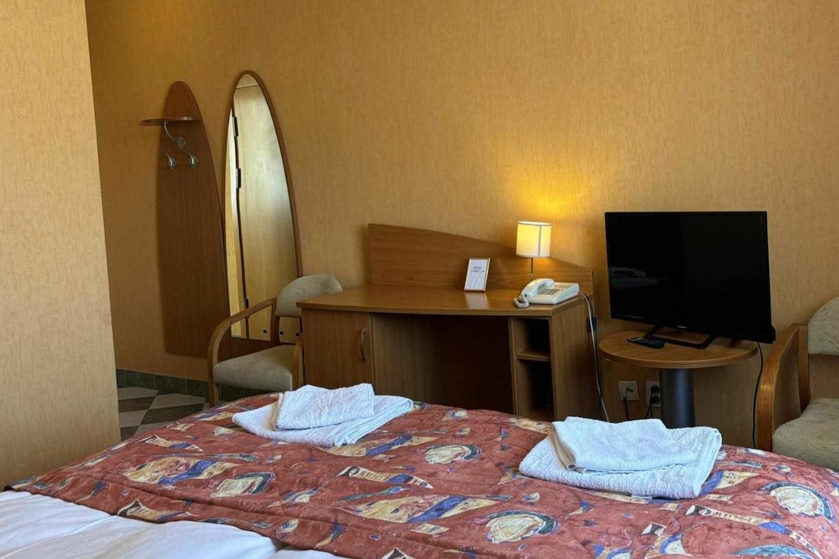 Habitación de hotel con cama, escritorio con teléfono y lámpara, televisión y una tabla de surf apoyada en la pared.