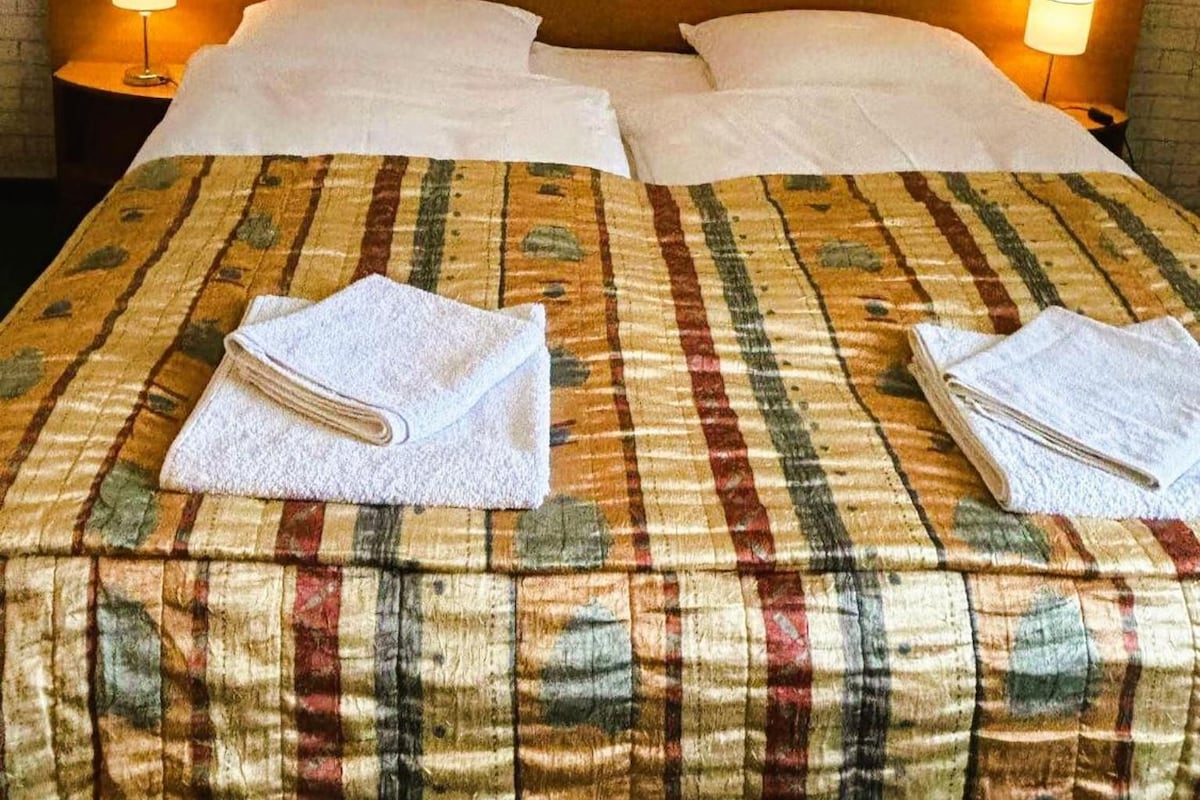 Habitación de hotel con pared de ladrillo, cabecero de madera, dos lámparas de noche, colcha a cuadros y dos toallas dobladas con cuidado.