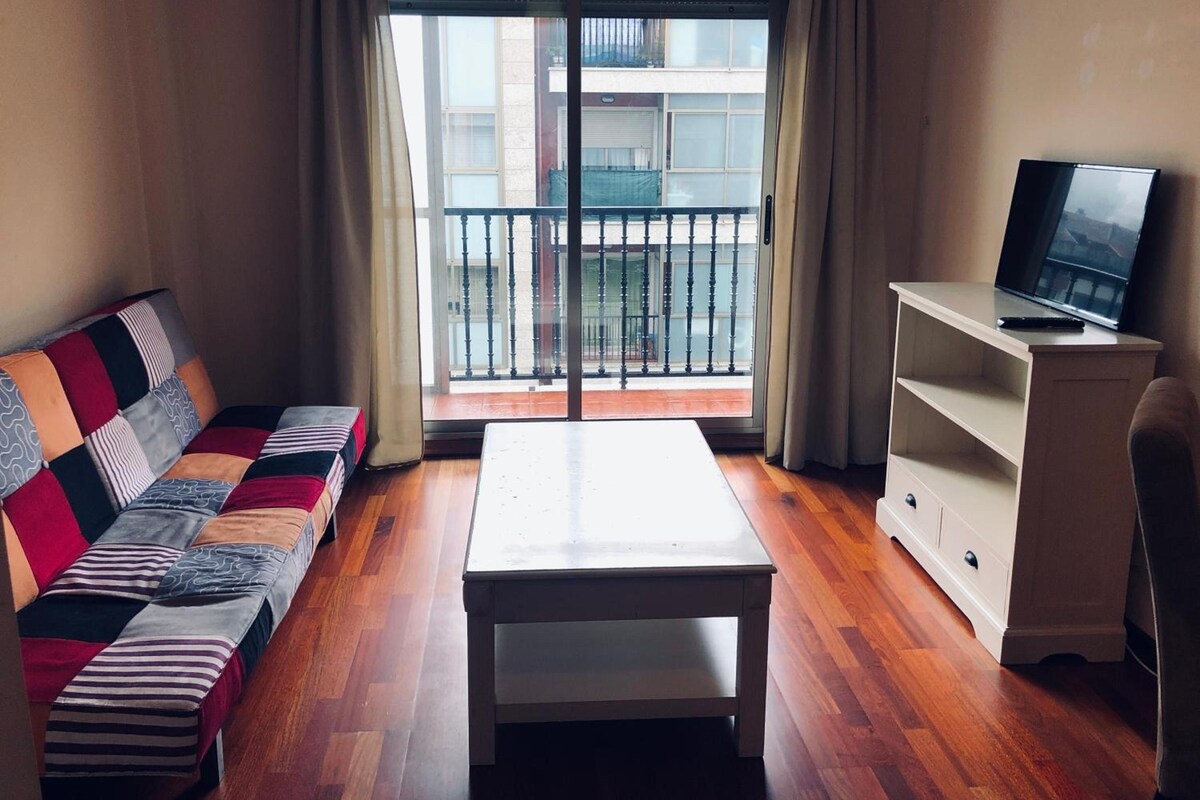 A room with a sofa, a table, a TV on a stand, and a view through a window.