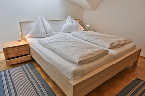 Un dormitorio moderno con cama de madera, ropa de cama blanca, una mesita de noche con cajones y una lámpara de noche.