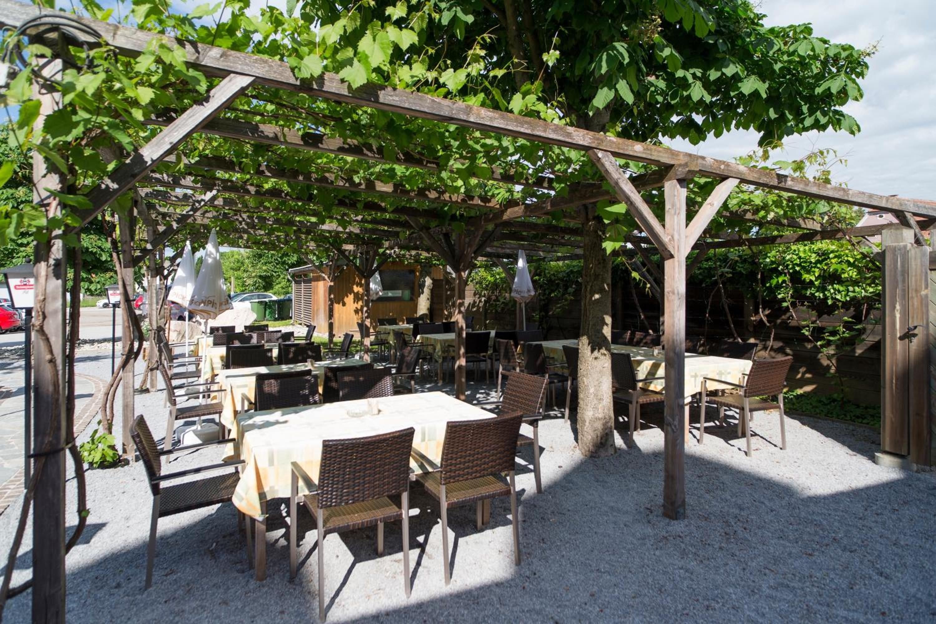 Ein Sitzbereich im Freien mit Tischen und Stühlen unter einer Pergola, umgeben von Grünflächen.