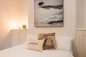 A bedroom with a bed, a pillow with 'WANDERER' text, a wall-mounted painting of a house by the sea, and a nightstand with a lamp and a plant. - The Terraces Mudgee (Mudgee)