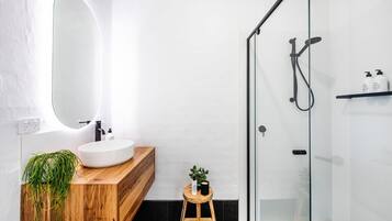 A modern bathroom with a wooden vanity, a round mirror, a small stool with a plant, and a glass-enclosed shower area.