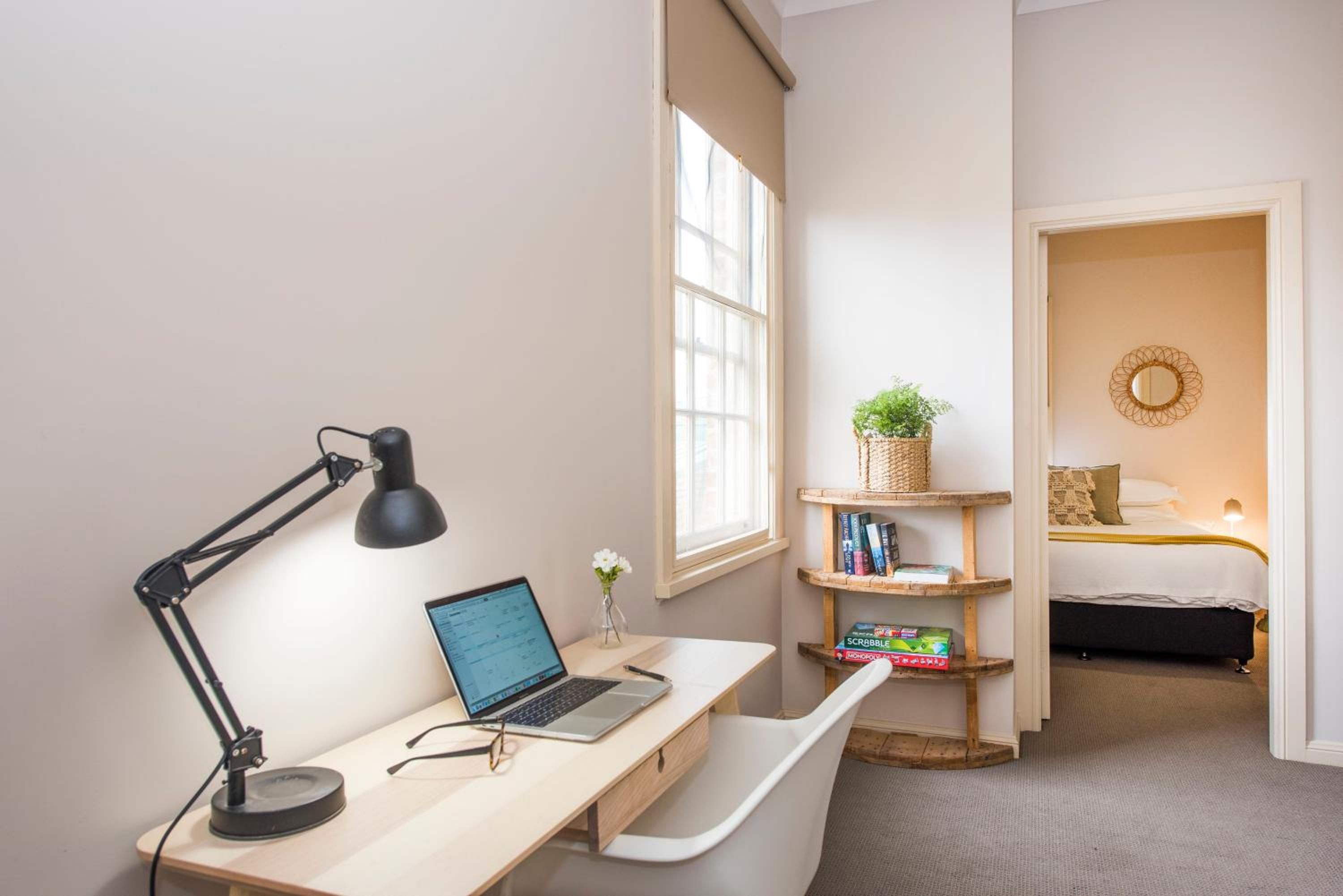 Una habitación con escritorio, computadora portátil y lámpara de pie. Hay una ventana con persianas, una estantería con libros y una planta, y una cama con cabecera y almohadas.