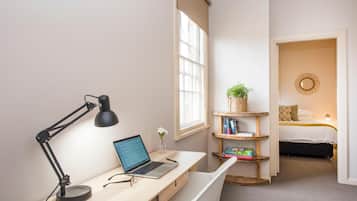 Una habitación con escritorio, computadora portátil y lámpara de pie. Hay una ventana con persianas, una estantería con libros y una planta, y una cama con cabecera y almohadas.