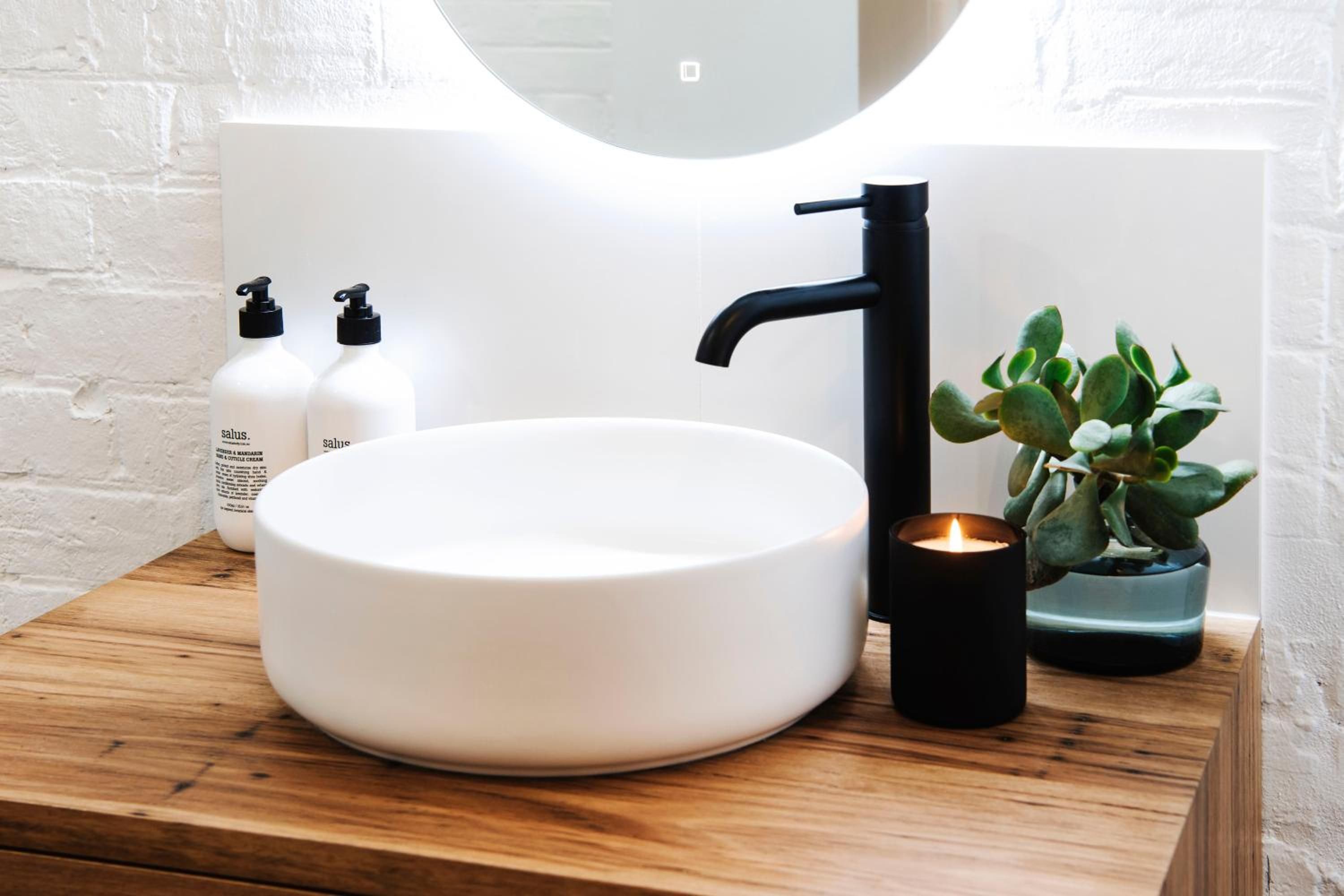 A bathroom with a white sink, black faucet, and a lit candle. There are two bottles of lotion and a potted plant.