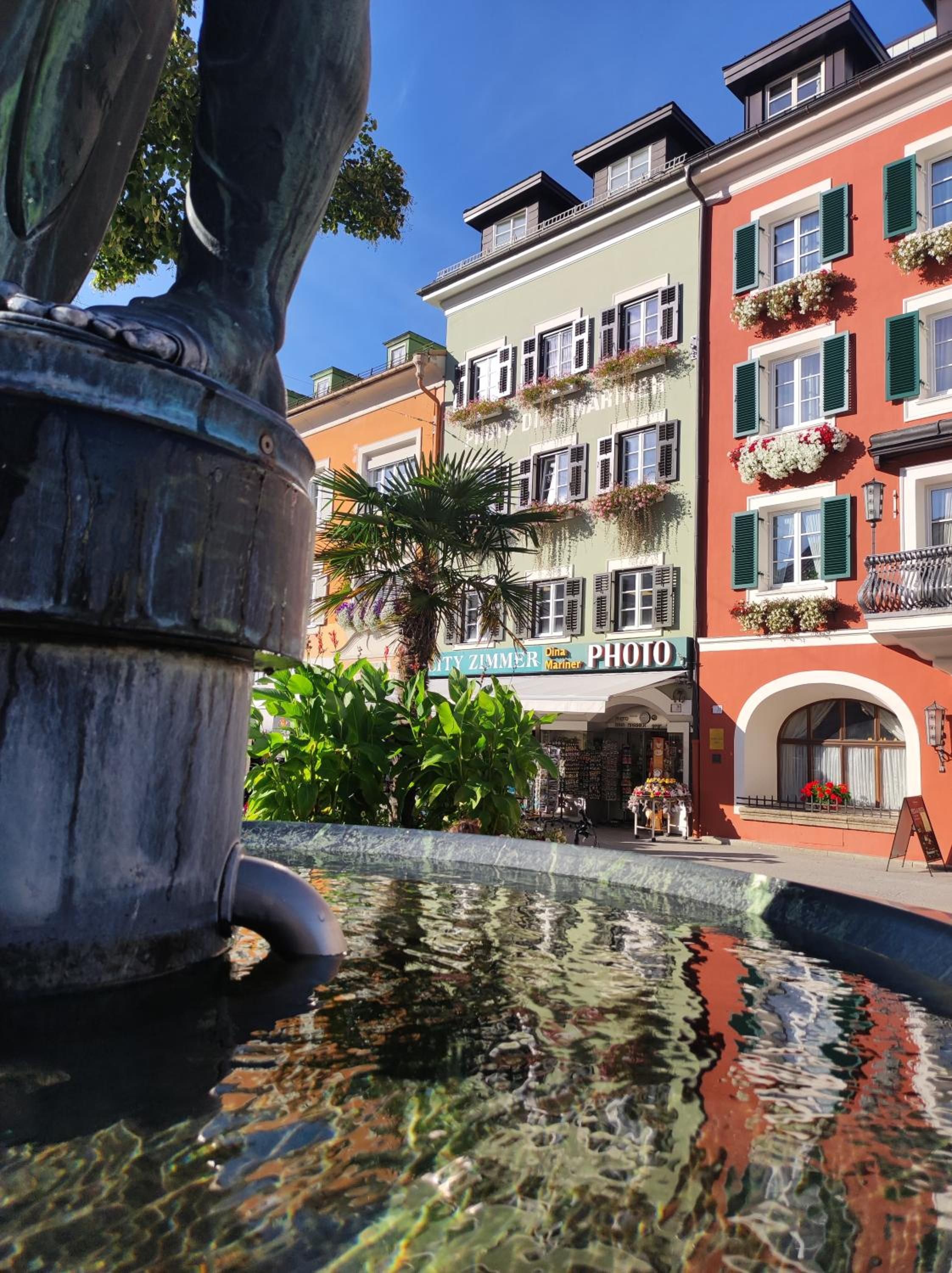 Ein Brunnen mit Statue, umgeben von Gebäuden mit Balkonen und Blumenkästen.