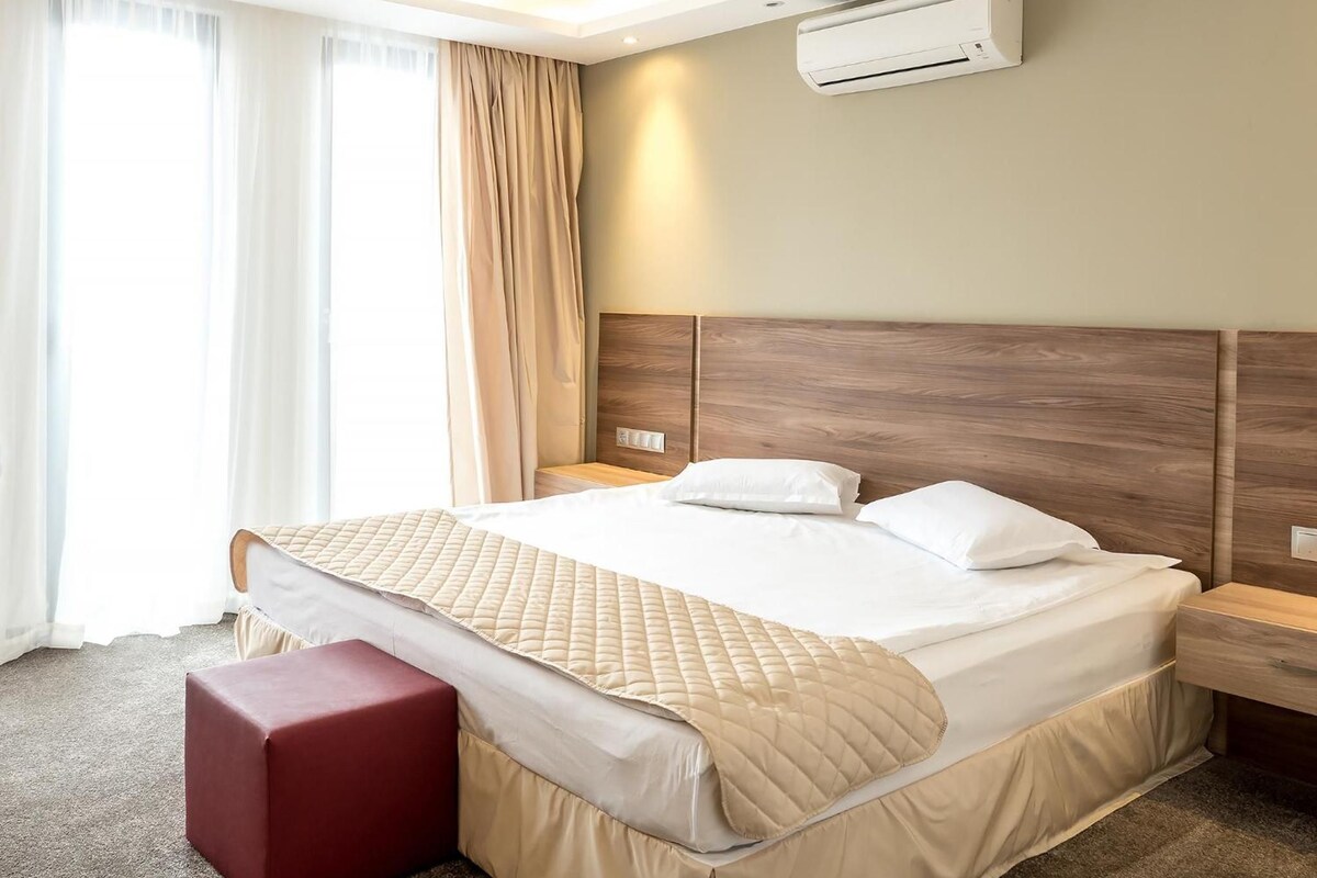 A hotel room with a large bed, a bedside table, an air conditioning unit, and a window with curtains.